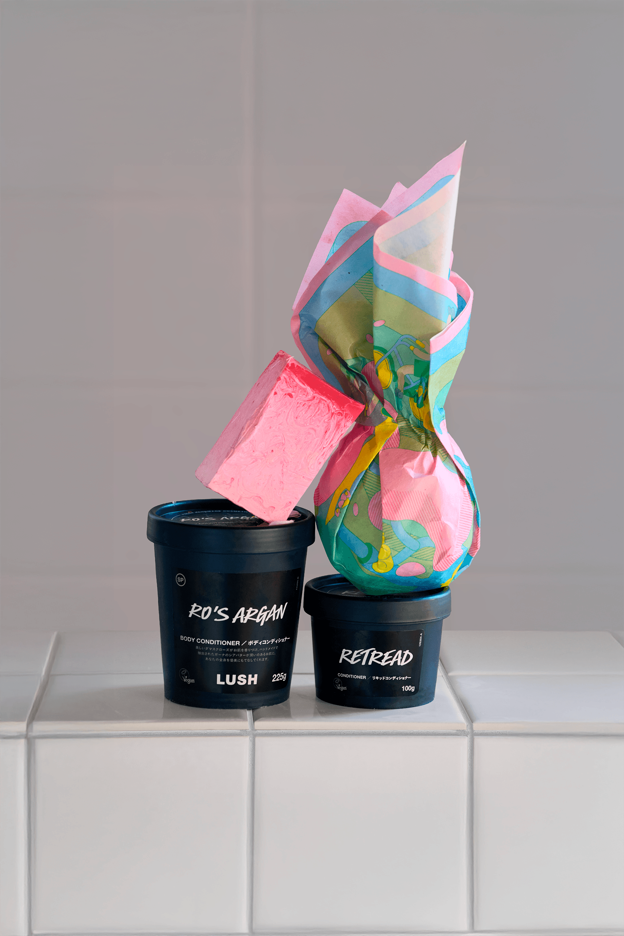 Wrapped bath bomb in translucent illustrated paper placed on top of black Lush product pots, displayed on white tiles.