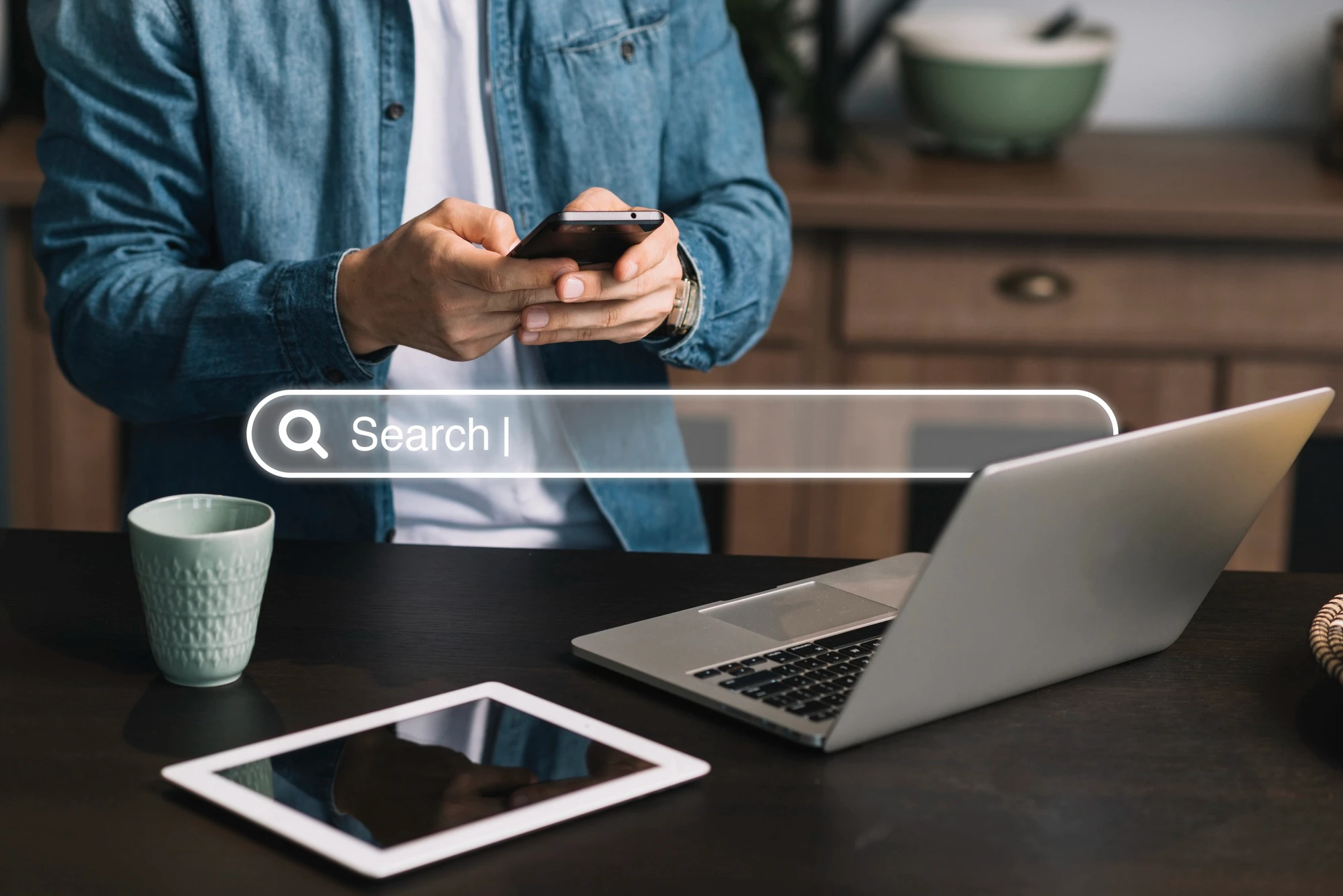 O corpo de um homem vestindo uma camisa jeans está de pé atrás de uma mesa preta com um laptop aberto, um tablet e uma xícara. Ele segura e olha para o seu smartphone. Uma barra de pesquisa flutua no ar, na altura de sua cintura, com uma lupa no canto esquerdo e a palavra "Search" escrita.