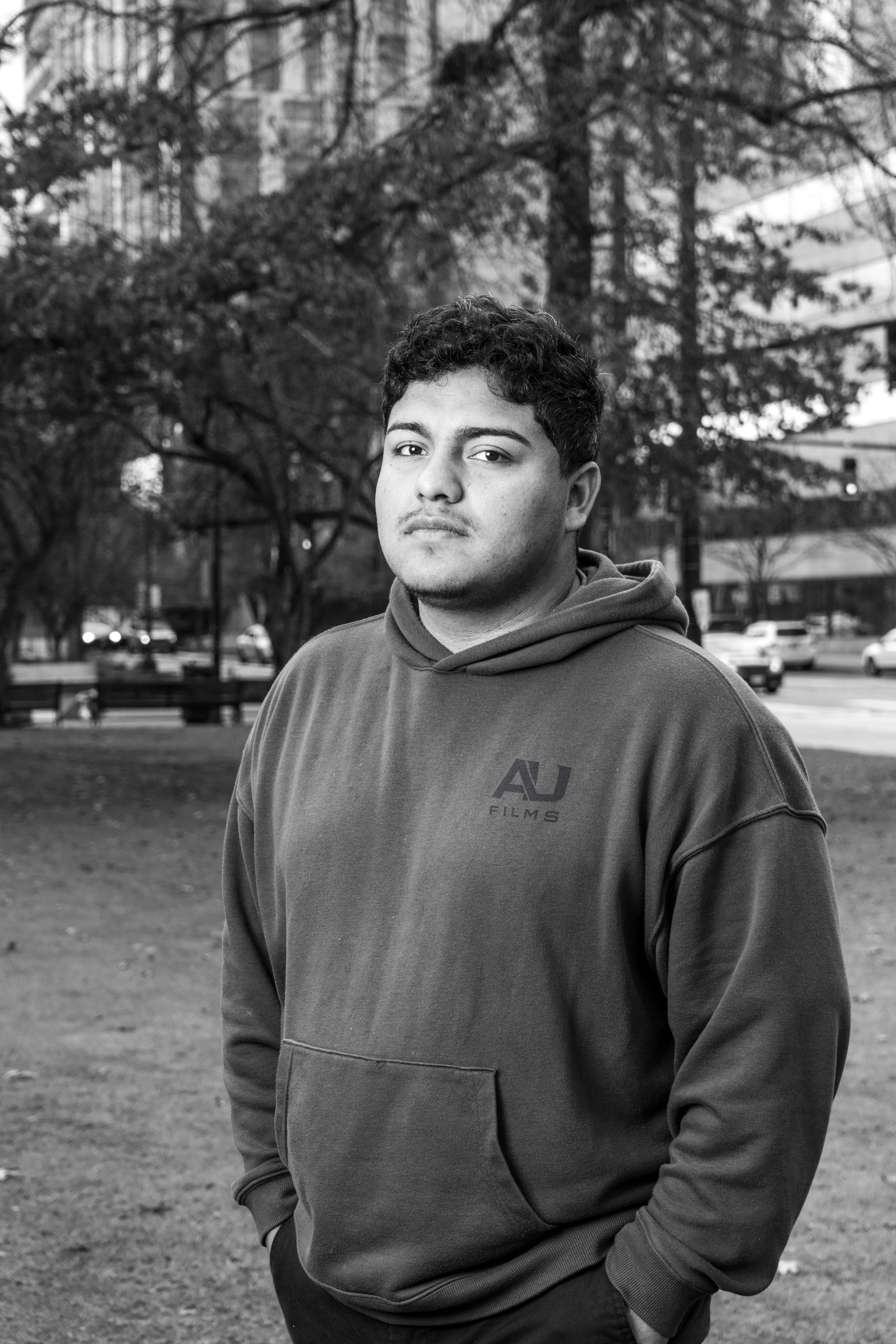 A groom in a dark suit carries a bride in a white dress across a sunny, grassy field. The couple shares a joyful, intimate moment with trees in the background.