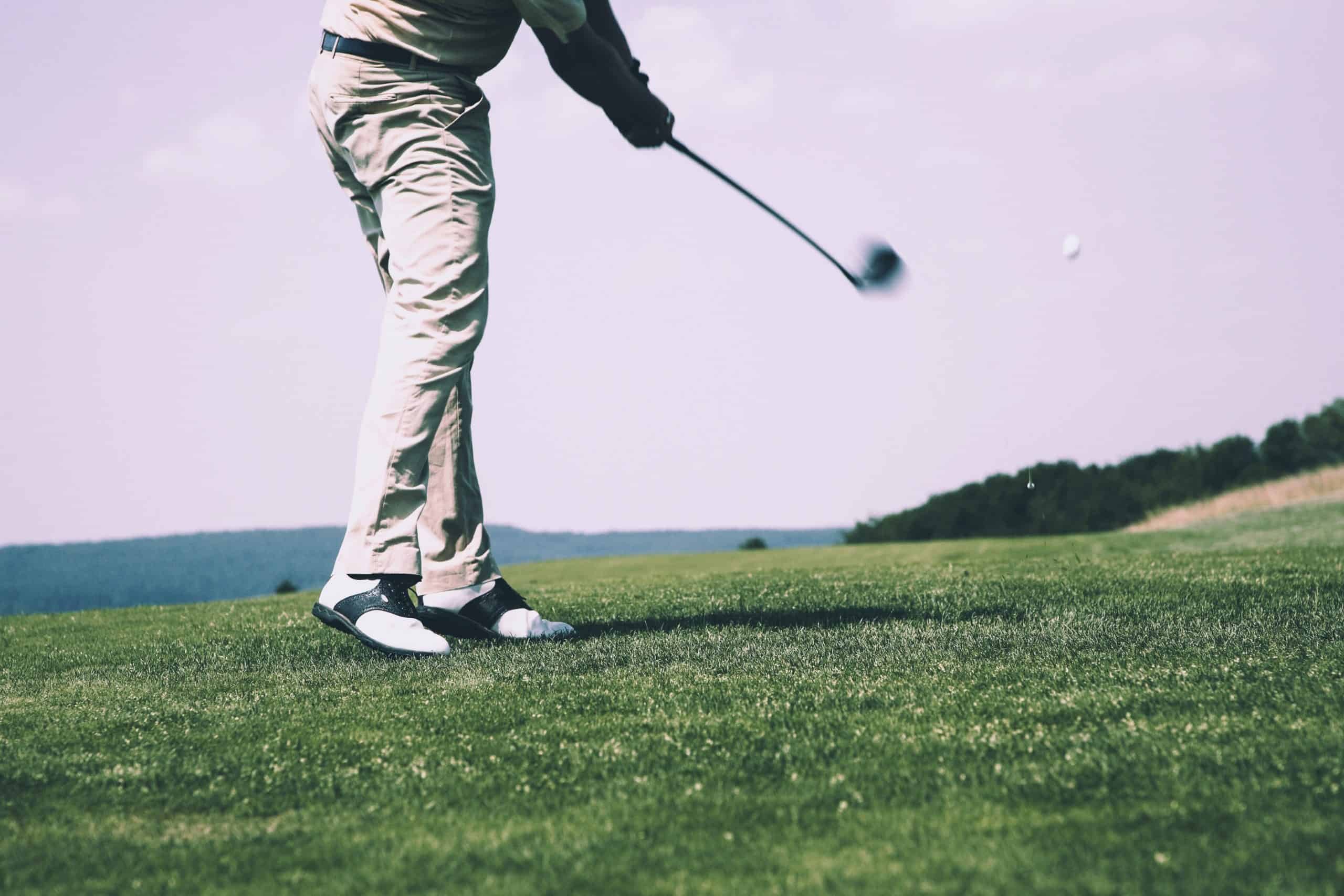 A golfer in mid-swing on a lush green fairway, with hills visible in the background, under a clear sky.