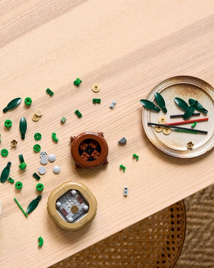 An assortment of small LEGO pieces, including green leaves, circular elements, and a brown container, spread across a wooden tabletop, accompanied by a decorative plate with additional pieces.