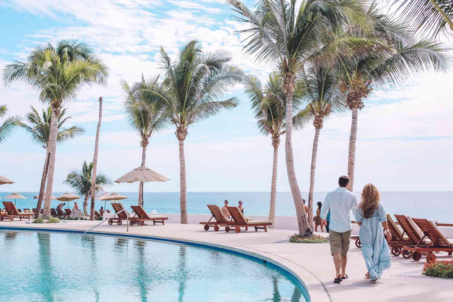 Couple strolls past the pool at an oceanfront all-inclusive resort in Mexico, framed by palm trees and the turquoise sea.