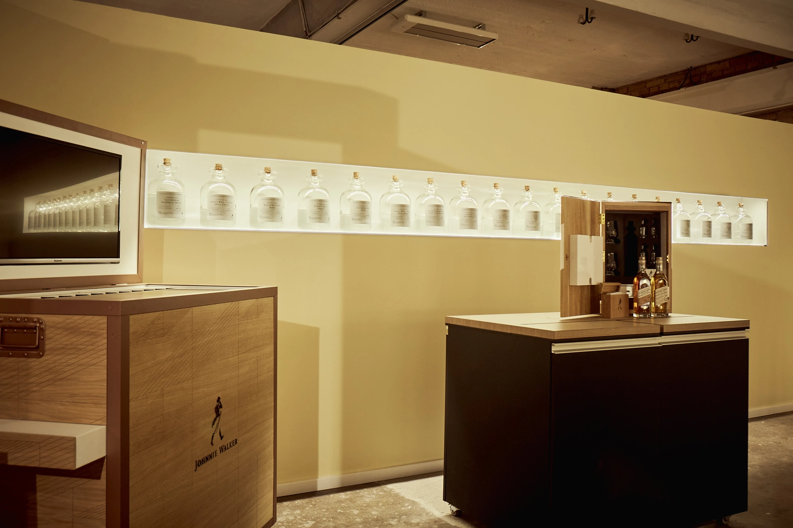 Backlit shelf with a long row of clear whisky bottles above a Johnnie Walker wooden crate counter at DIAGEO BCB17.