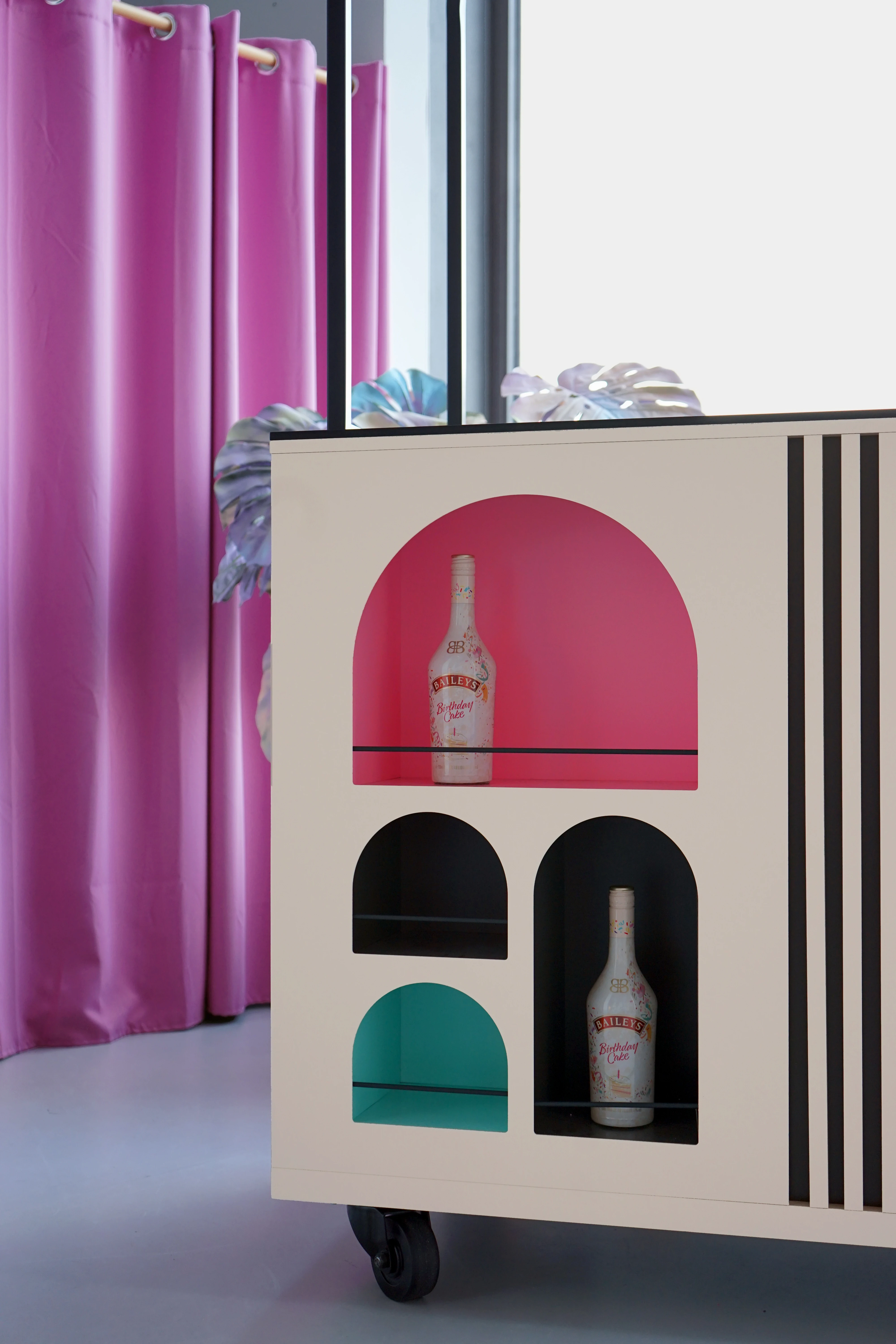 Detail of the BAILEYS Treatbar body with arched niches in pink, black and turquoise holding Baileys bottles, on castors in front of a pink curtain.