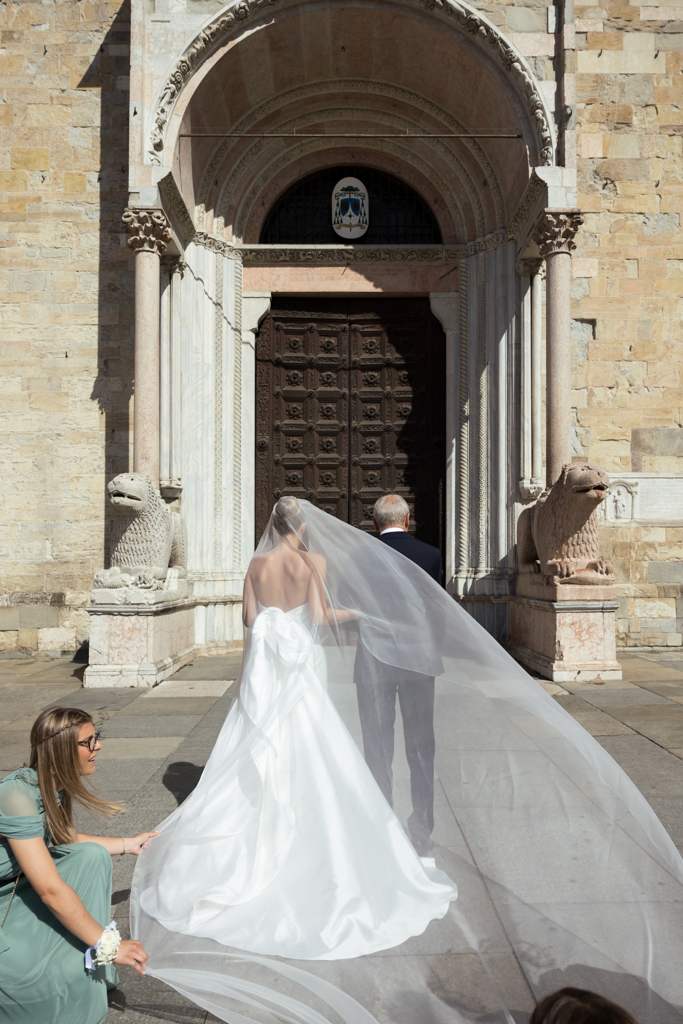 Sposa e velo lungo davanti al portale barocco di Villa Malenchini in reportage matrimoniale, assistente regge lo strascico