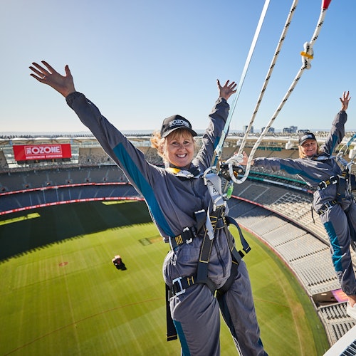 Two people in harnesses with raised arms smile while standing on a stadium roof, with empty stadium seats below them.