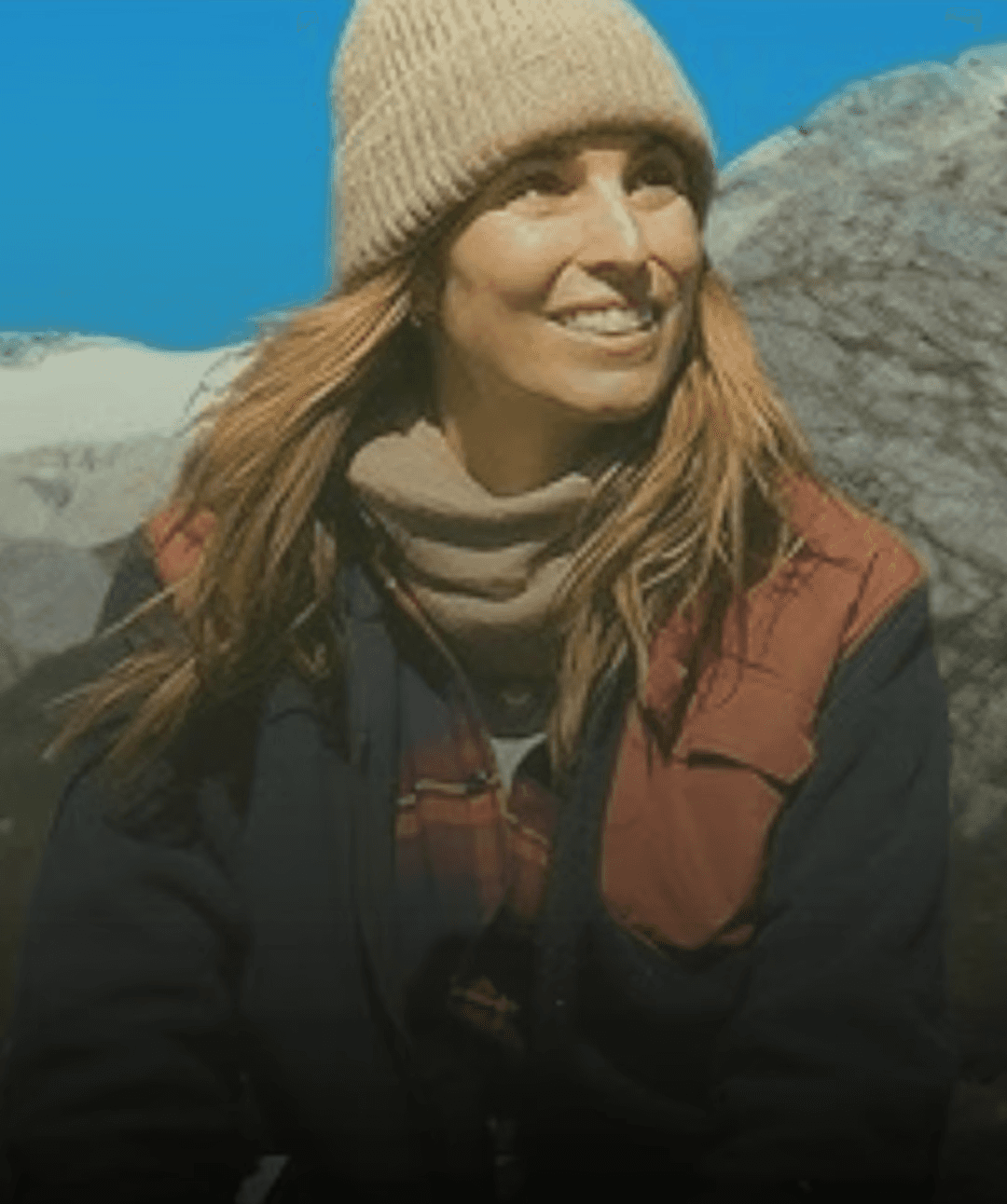 A smiling woman with curly hair is posing in a warmly lit indoor setting. She wears a dark-colored shirt.