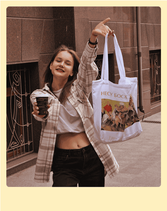 дівчина стоїть на вулиці і тримає в одній руці каву у іншій кастомну еко-сумку з надрукованим малюнком