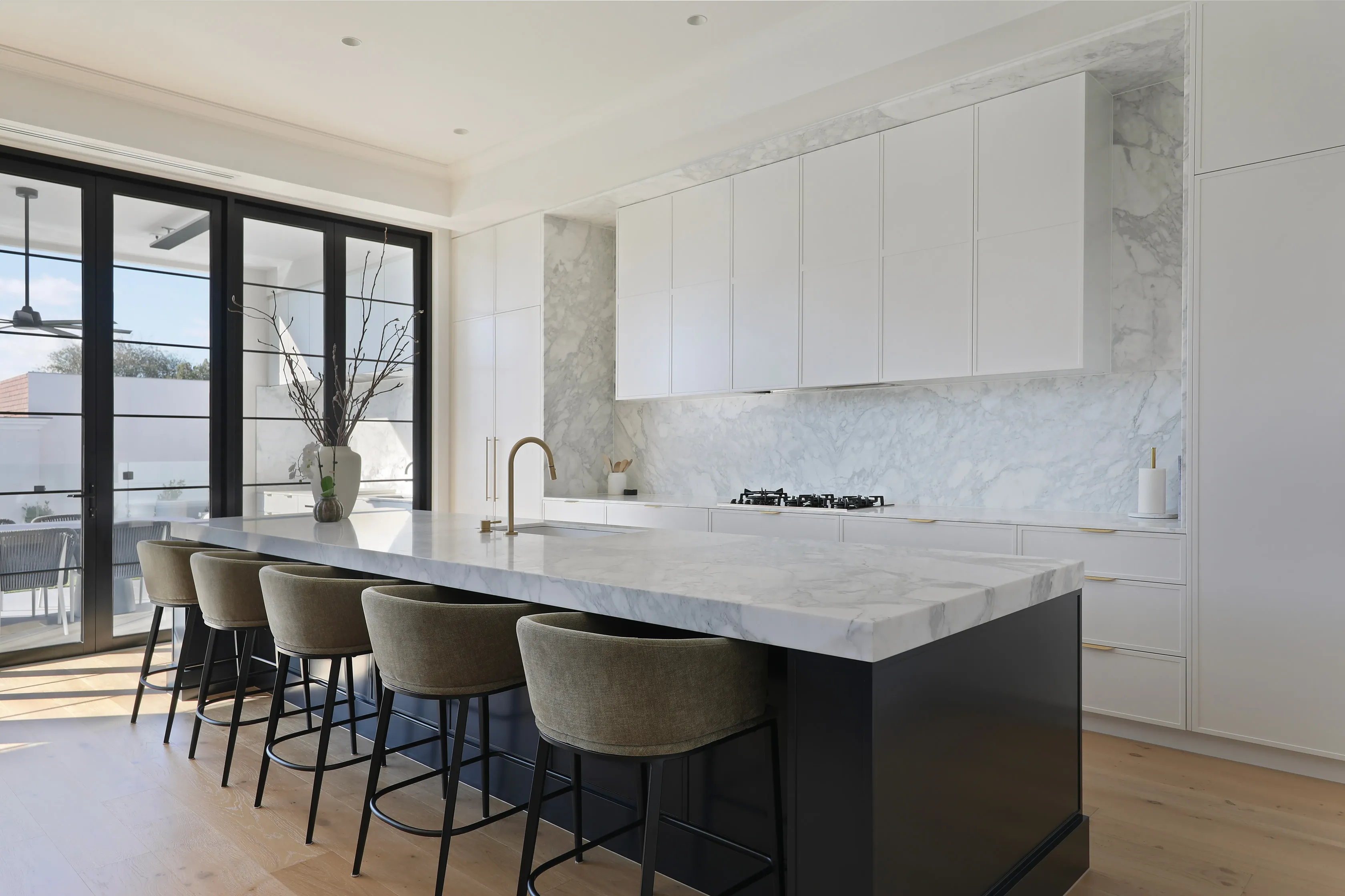 Kitchen with white cabinetry, a Calacatta marble splashback and waterfall island with brass gooseneck tap, olive bar stools and black-framed doors to a sunlit terrace
