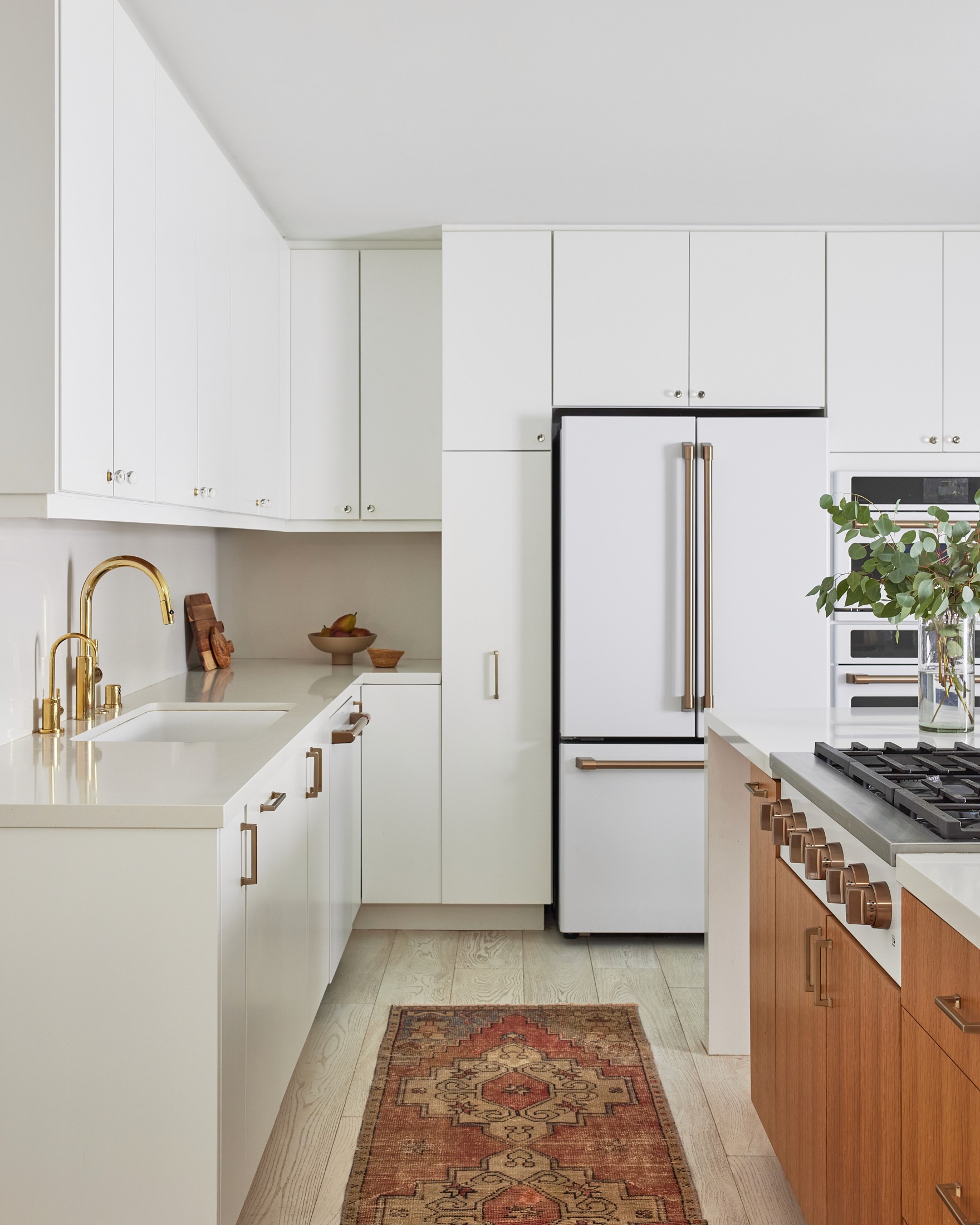 White kitchen with bronze fixtures, white cabinets, refrigerator with coordinating panels, bright white quartz countertops, island range, Turkish rug, and warm wood accents by Jubilee Interiors in Los Angeles, California