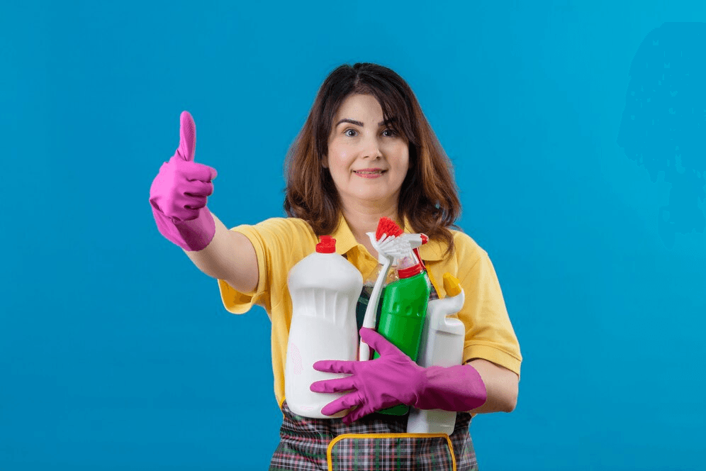 Woman cleaning enthusiast giving a thumbs-up while holding several cleaning bottles, wearing pink gloves and a yellow shirt, in front of a blue background.