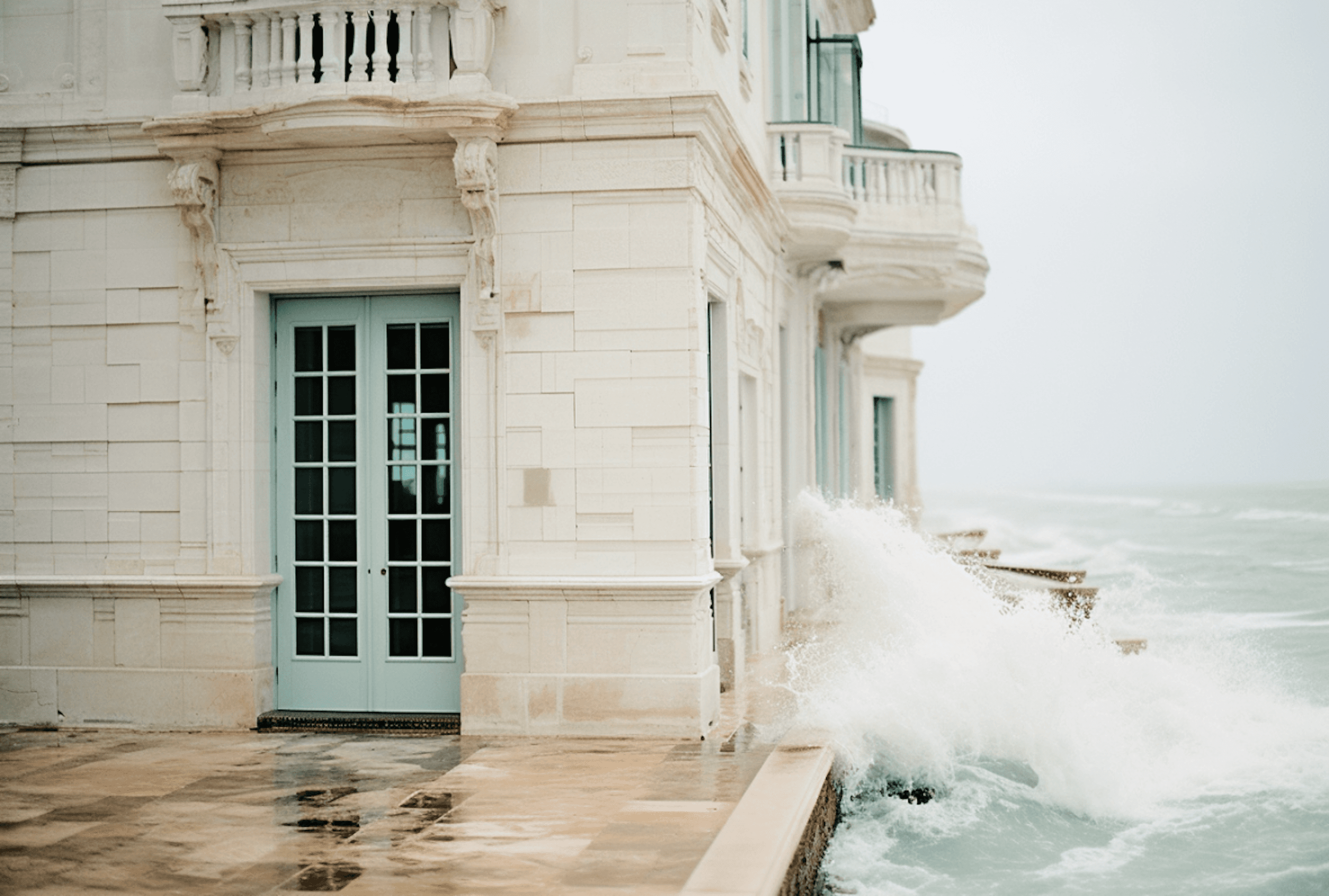 A white seaside hotel with balconies and green-framed doors above waves breaking on the rocks