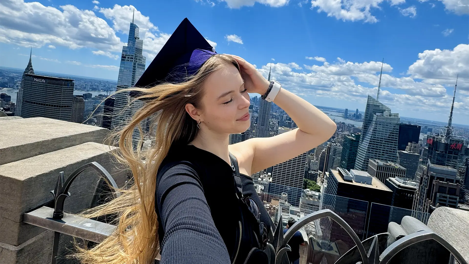 Studentka Nina v akademické čepici na vyhlídce v New Yorku s panoramatem mrakodrapů v pozadí.