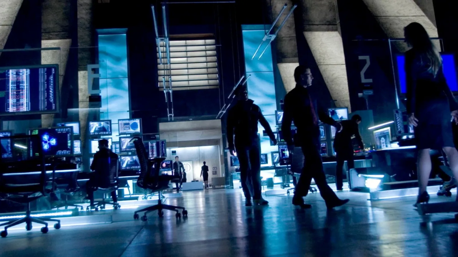 A modern, high-tech control room with numerous computer monitors displaying data, blue ambient lighting, and people in business attire actively engaged in discussions and operations.