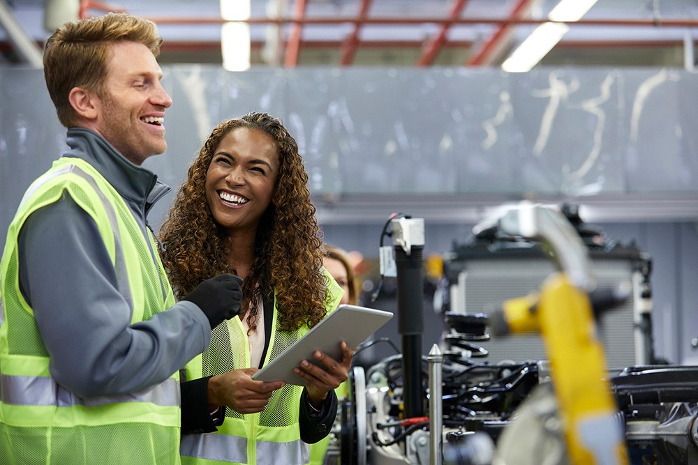 Two workers in hi-vis vests laughing together on a factory floor, one holding a tablet.