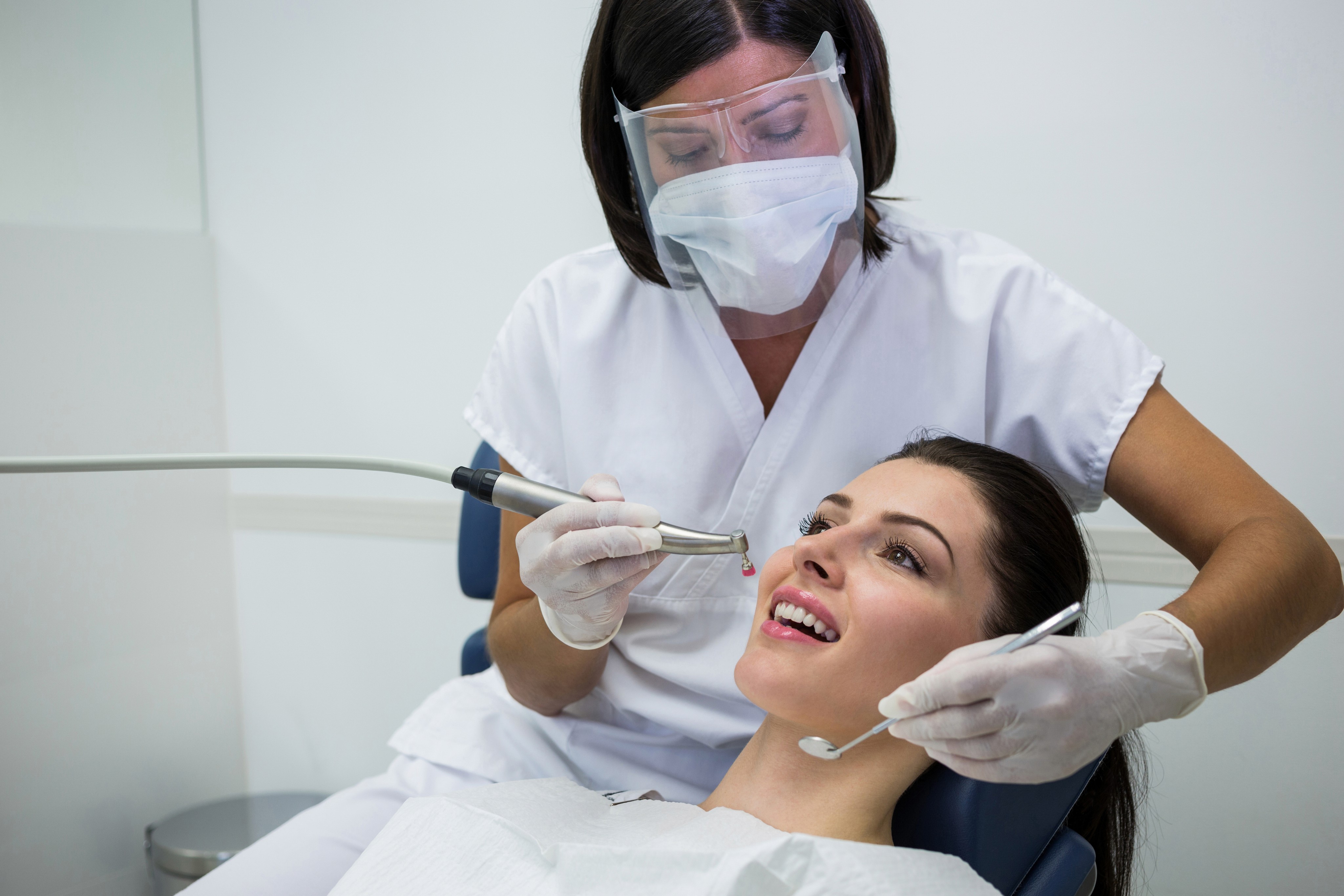 A dentist wearing a mask and gloves performing a dental procedure on a female patient who is smiling in the dental chair.