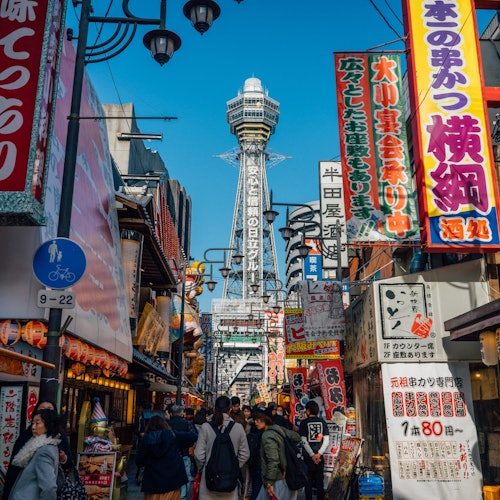 繁忙的日本街景，人群熙熙攘攘，色彩缤纷的广告牌，商店，以及在晴朗的蓝天背景下耸立的高塔。