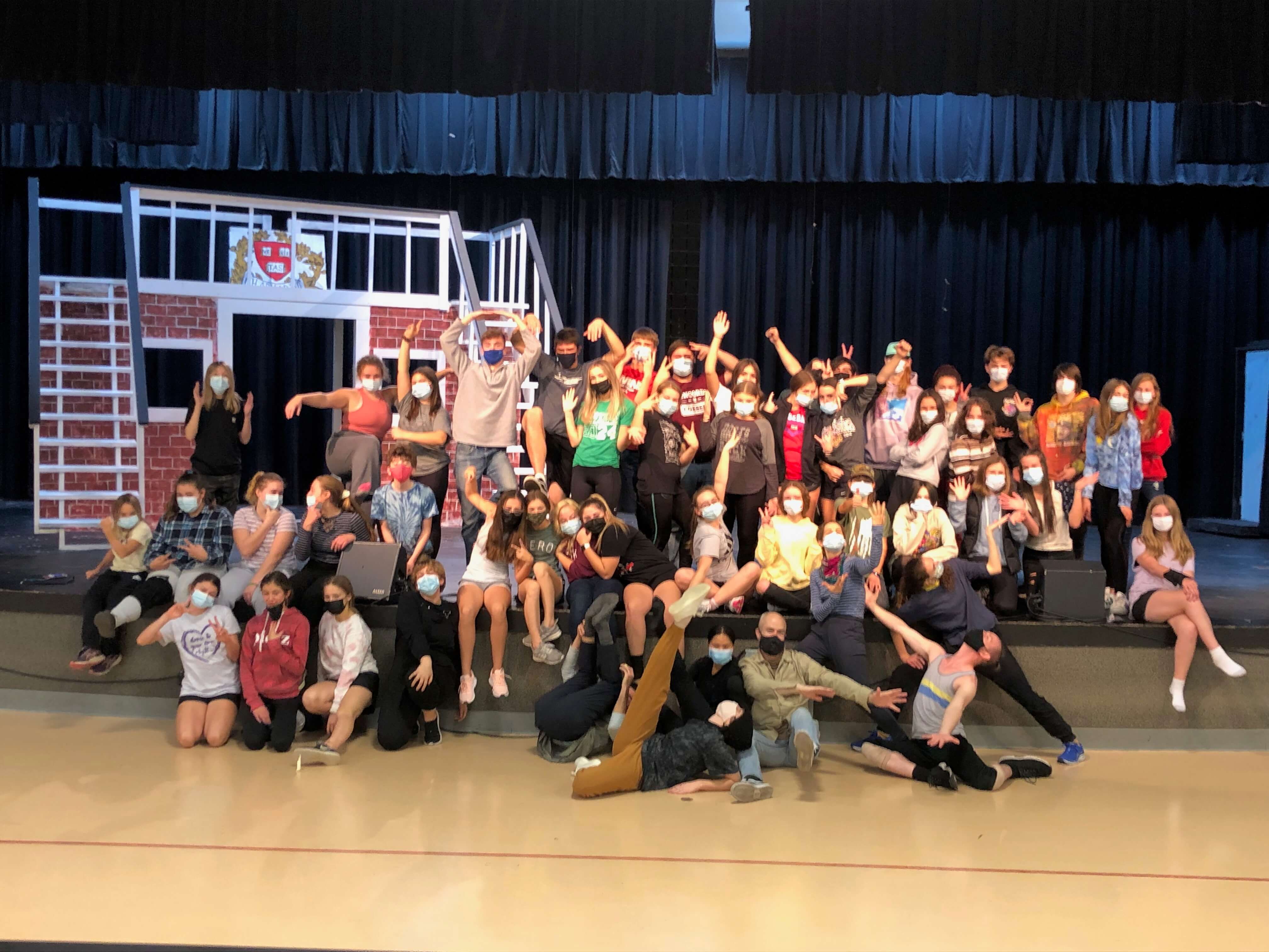 A large group of youth residency students in face masks gather together to pose happily on a theater stage floor.