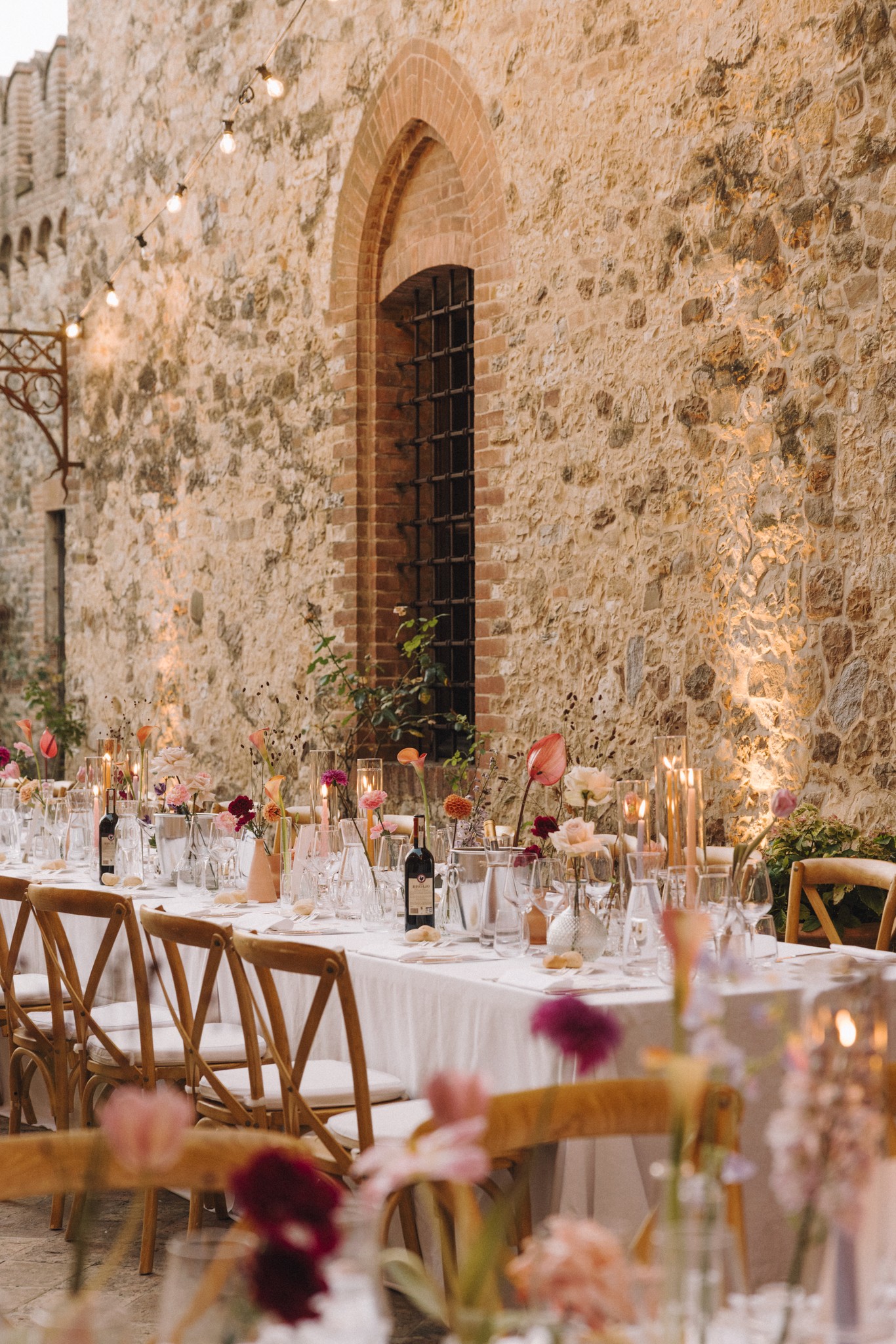 Tavolo ricevimento matrimonio al Castello di Tabiano con fiori rosa, sedie vintage e muro in pietra medievale sullo sfondo