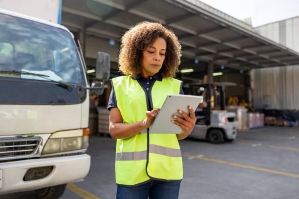 Woman wearing a hugh vis vest looking at a tablet stood in from of a lorry docked in a warehouse