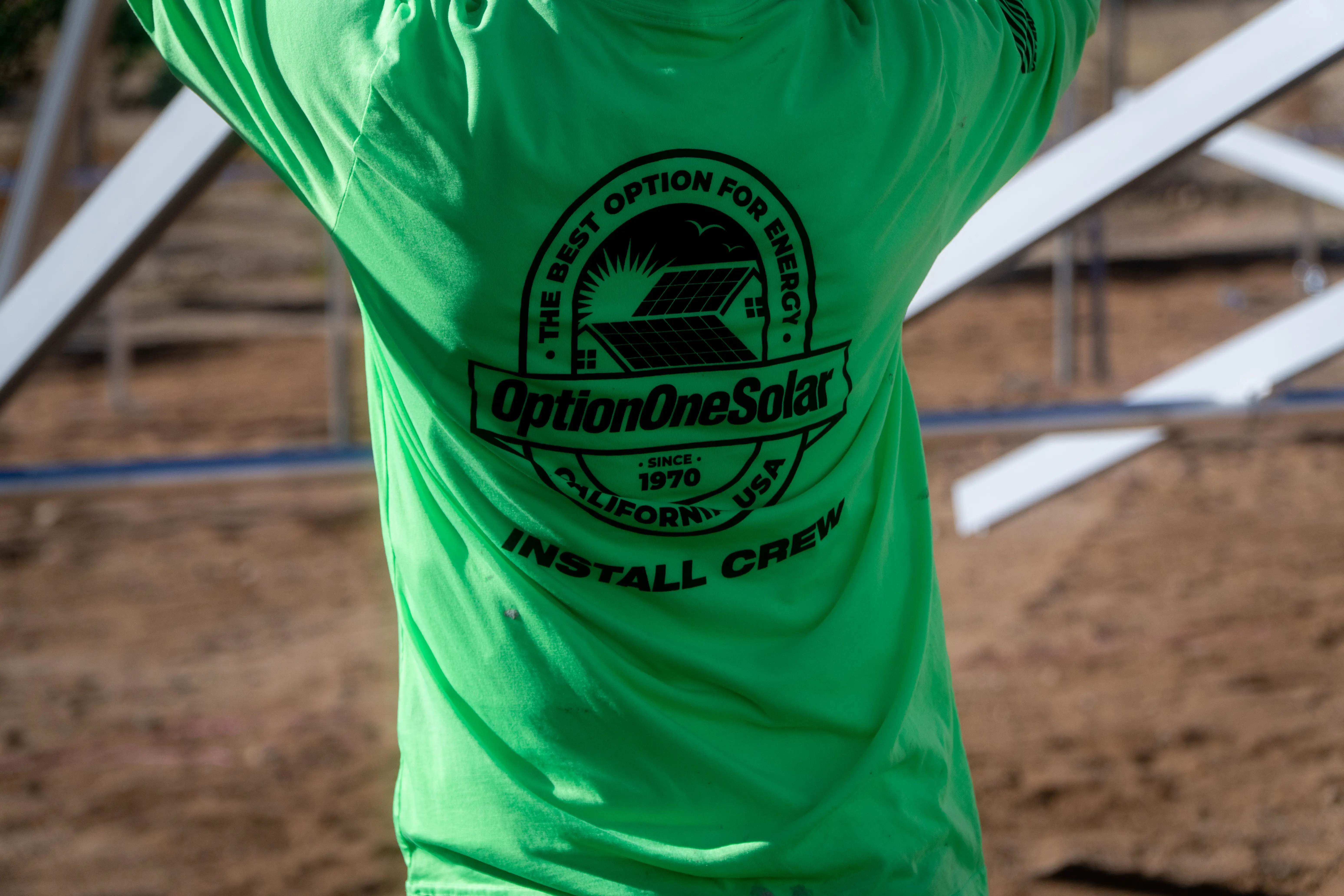 The back of a bright green Option One Solar install-crew shirt, printed with the company's since-1970 badge, at a ground-mount job site
