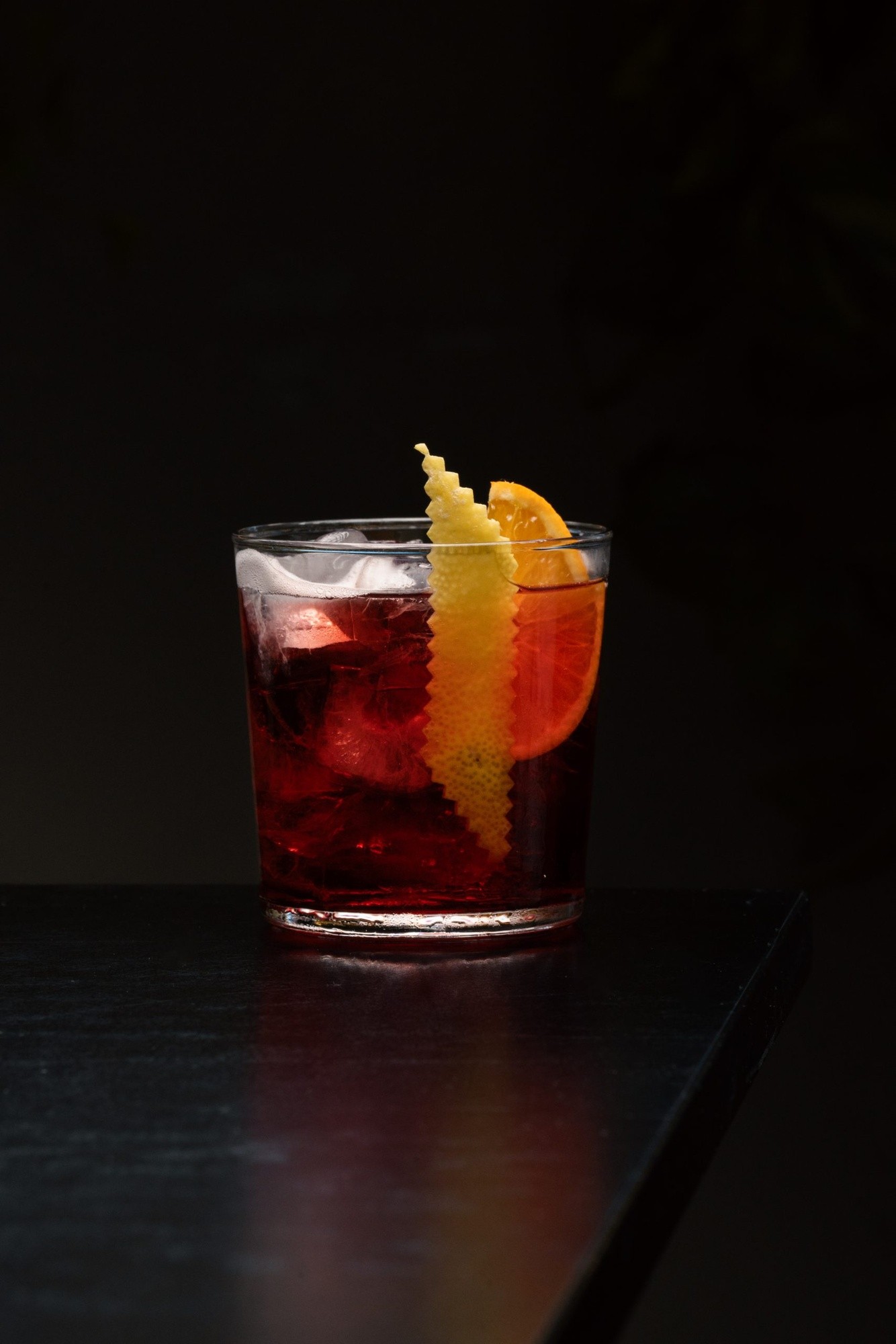 Cocktail rosso scuro servito in tumbler con guarnizione scorza e fetta arancia, su tavolo nero