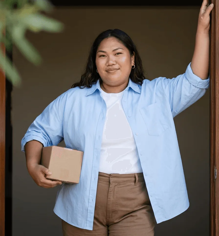 A smiling woman in a light blue shirt leaning in a doorway, holding a small parcel