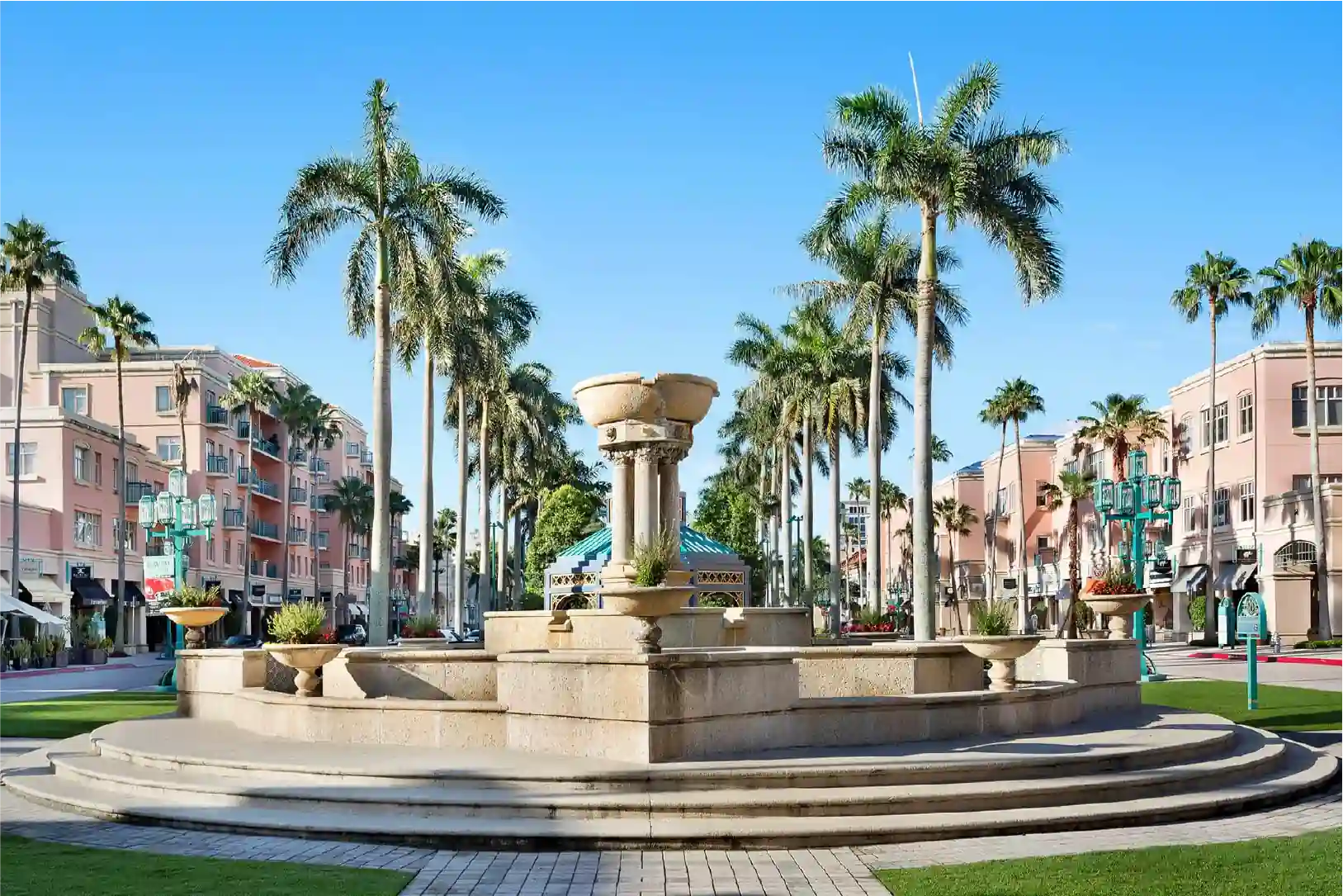 A grand stone fountain surrounded by palm trees in a lively plaza, with pastel-colored Mediterranean-style buildings lining the streets. The scene captures the vibrant and upscale atmosphere of Boca Raton, Florida, with boutique shops, cafes, and lush greenery under a bright blue sky.