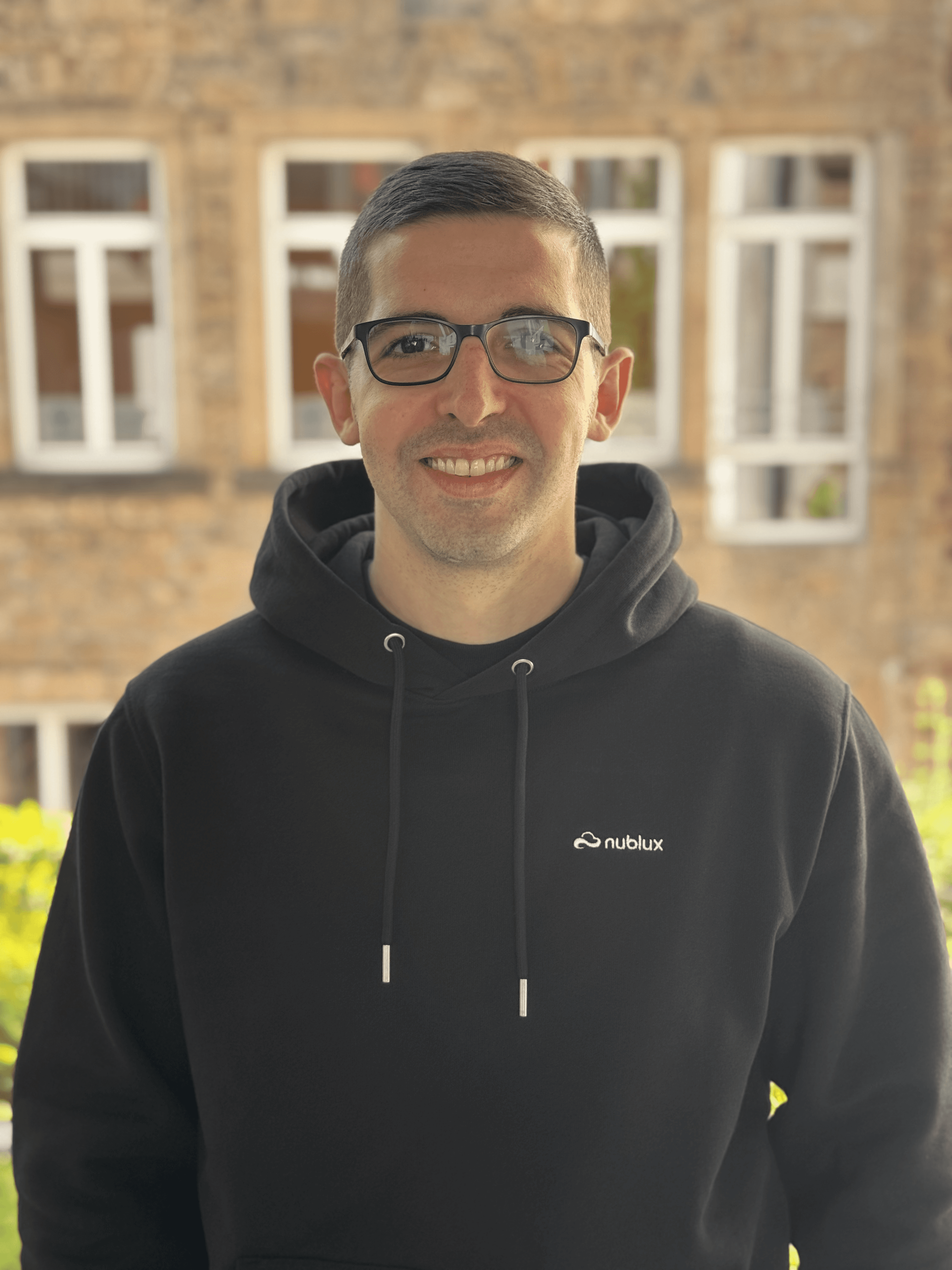 A smiling young man with crossed arms, wearing a plaid shirt and white t-shirt, poses against a dark background.