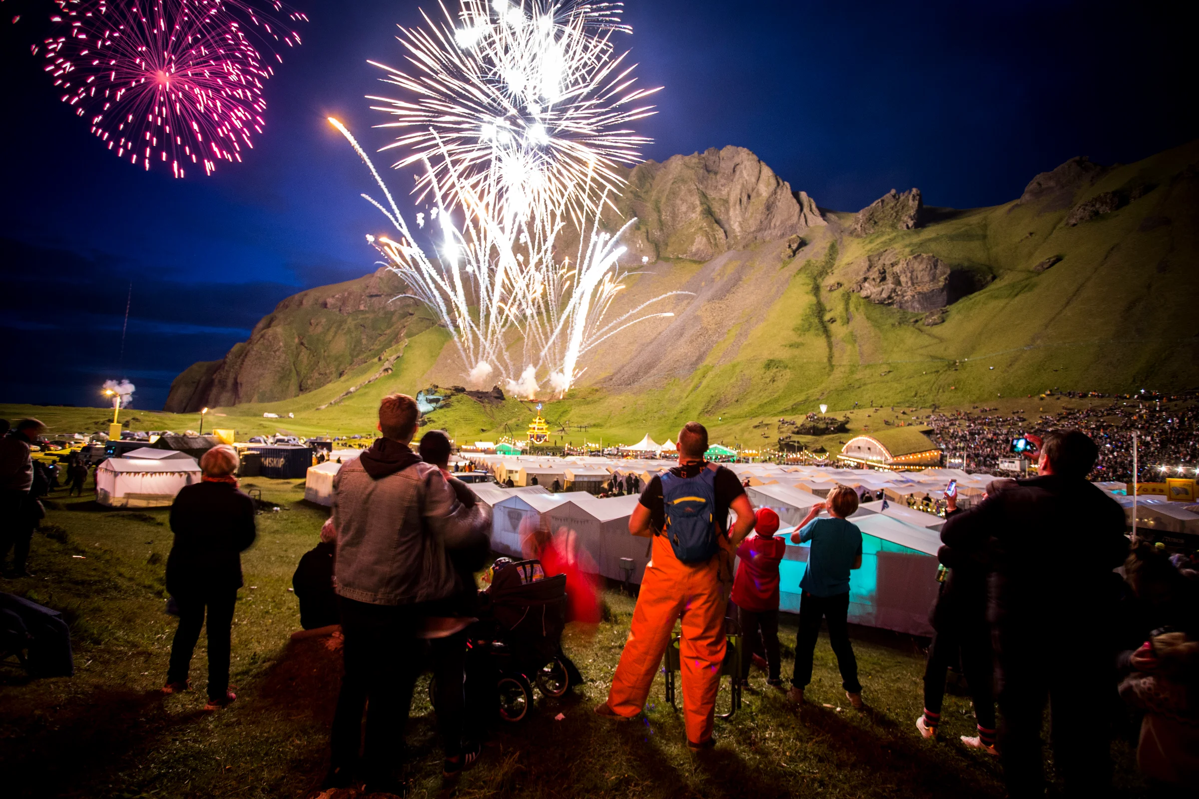 Spectators watch a bright fireworks display over rows of white festival tents in Herjólfsdalur Valley on Heimaey Island in the Westman Islands as part of Verslunarmannahelgi with a large volcanic hill in the background.