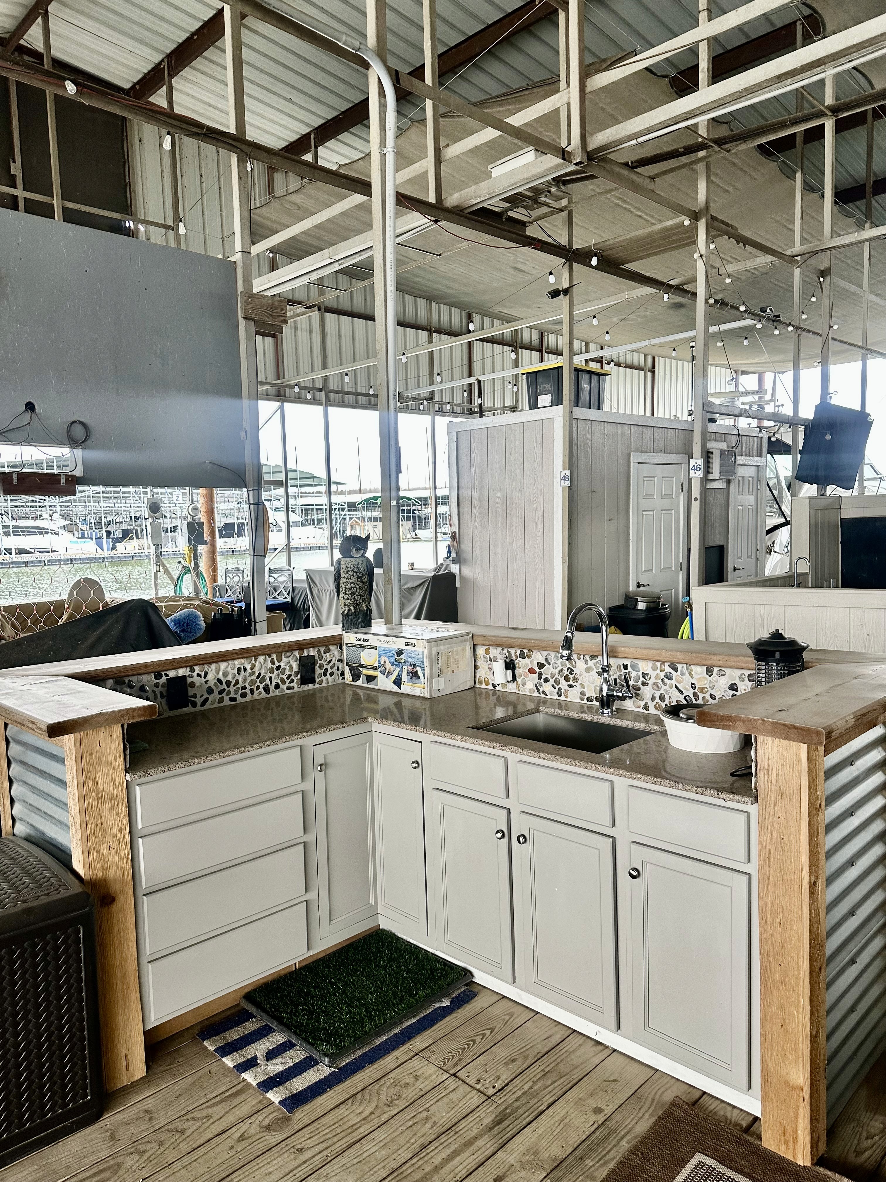 Outdoor kitchen with rustic wood accents featuring a granite countertop, mosaic tile backsplash, stainless steel sink, and white cabinetry, set under a corrugated metal roof with strings of lights in a spacious patio area.