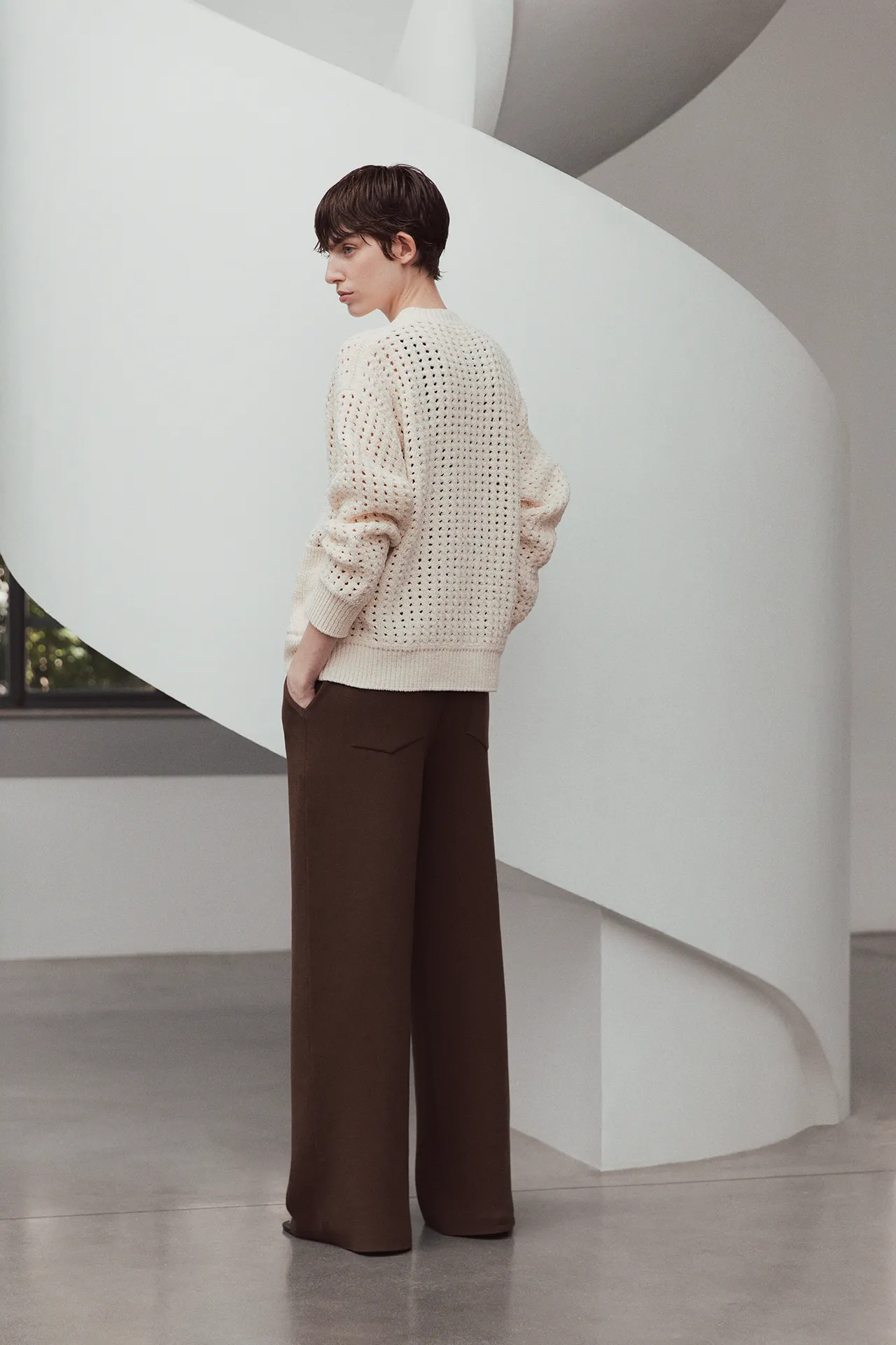 Rear view of a model from the Aida Barni SS26 collection wearing a cream chunky open-knit cardigan and brown wide-leg trousers standing near a white staircase.