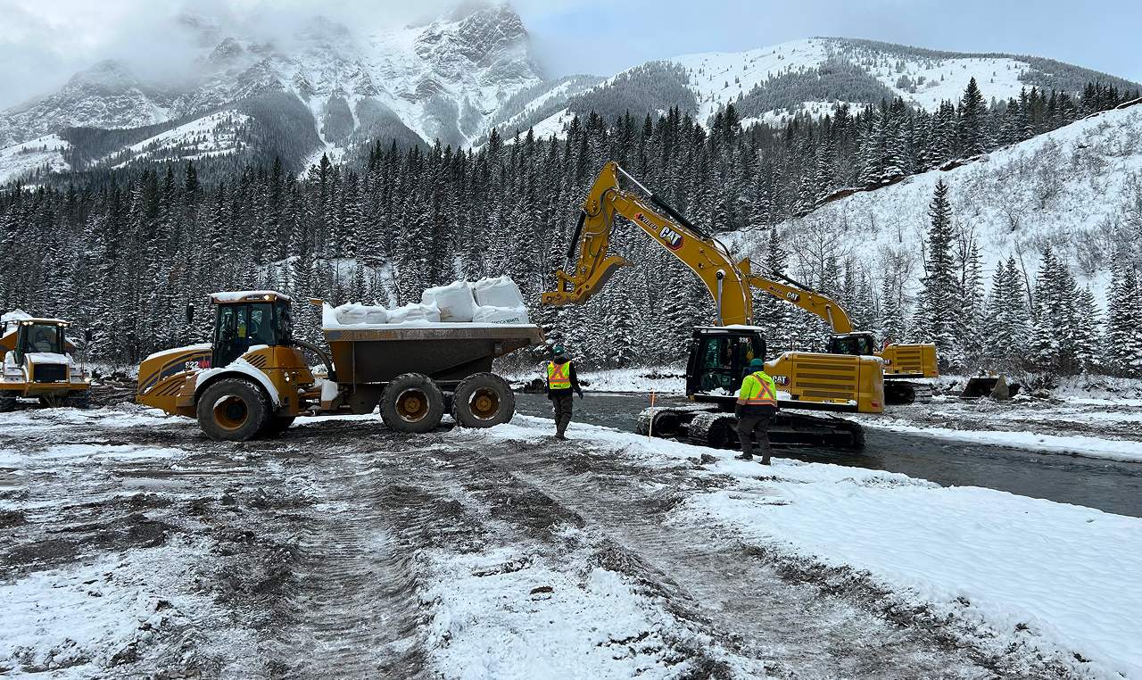 Winter sediment removal operations with excavator loading dump truck on Kananaskis River under snowy conditions