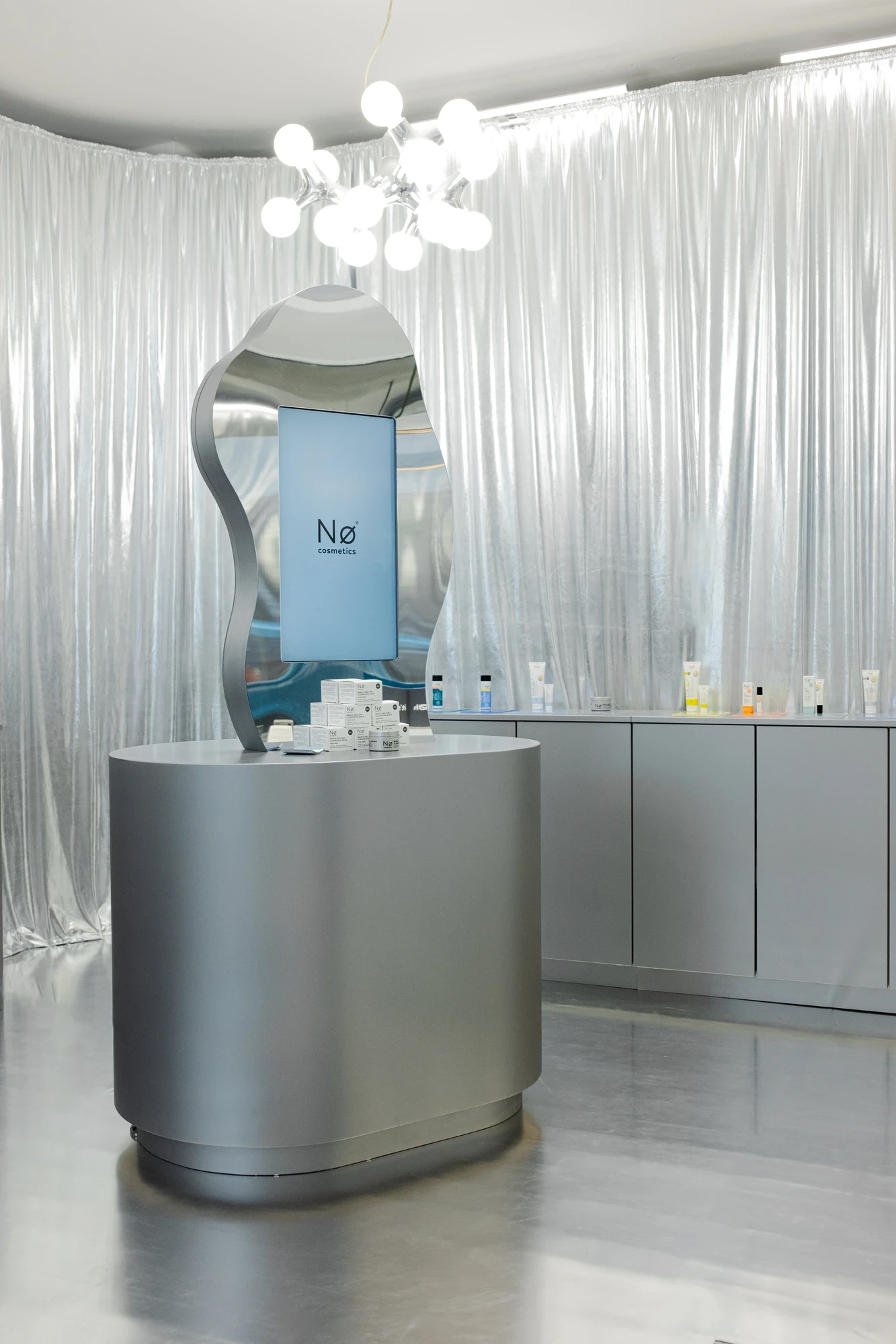 Nø Cosmetics booth with an oval metallic counter, sculptural chrome mirror, chandelier and silver curtain walls.