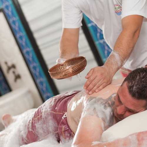 En person får en skummassage medan hen ligger med ansiktet neråt. En annan person häller vatten från en kopparskål över mottagarens rygg.