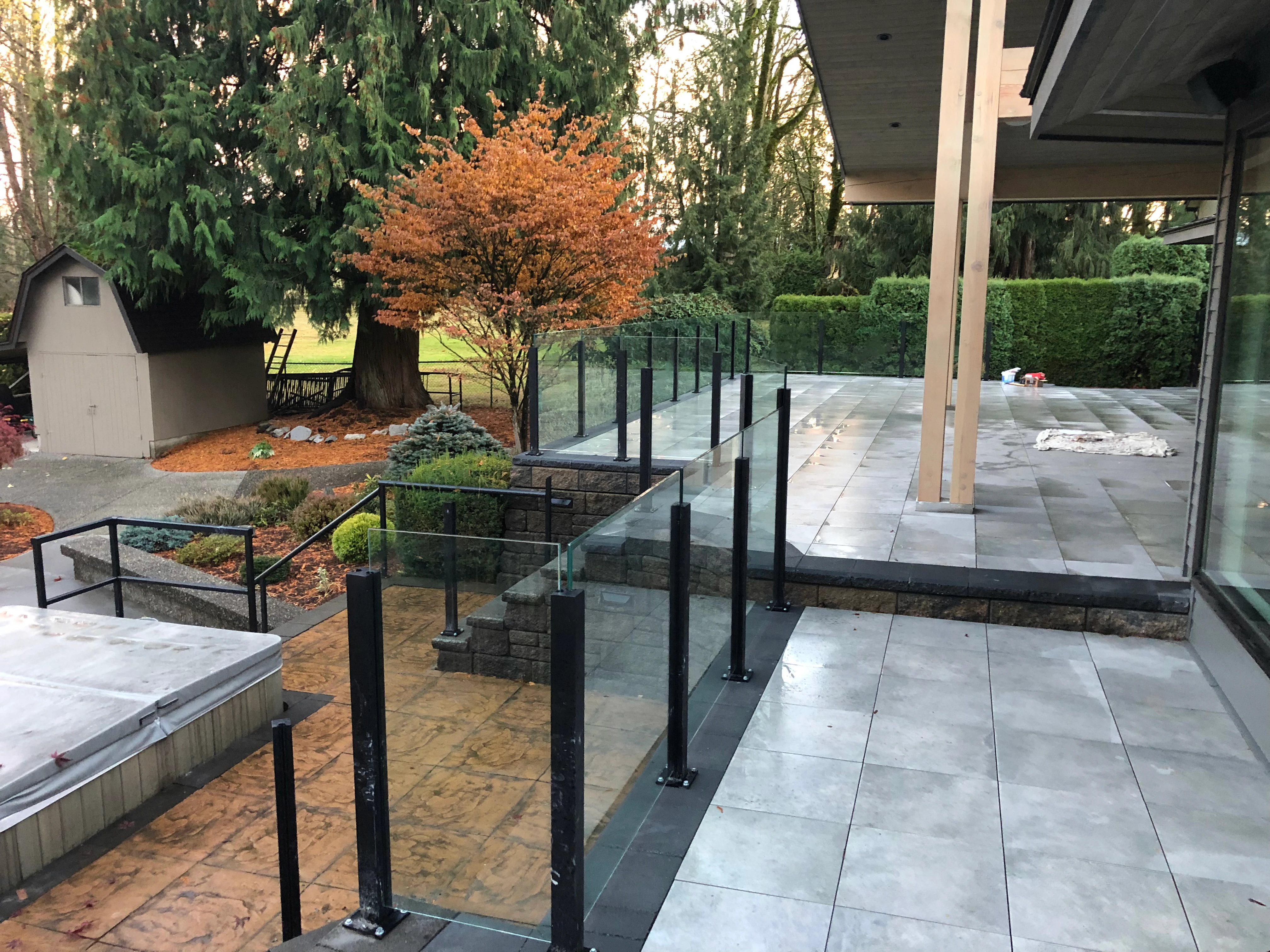 Modern outdoor patio and terraced backyard with large gray stone tiles, black-framed glass railings, and landscaped garden beds.
