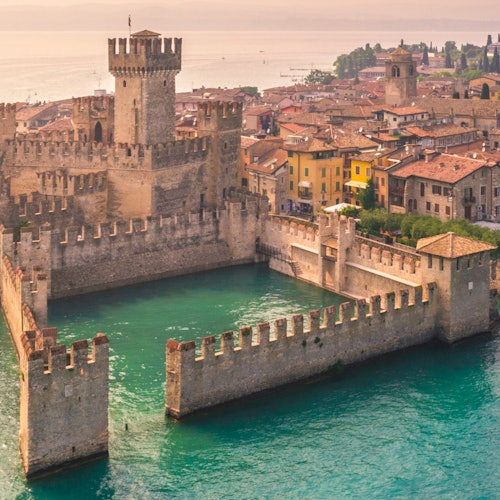 Aerial view of a historic stone castle with a square moat surrounded by a town with colorful buildings near a body of water.