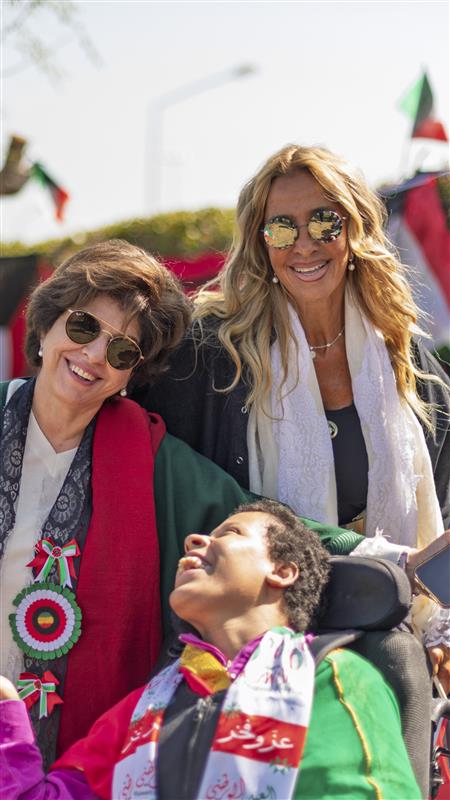 Una persona con una camiseta negra y pantalones de color claro está de pie en la cima rocosa de una montaña al atardecer, sosteniendo una cámara frente a su cara para tomar una foto. Un mar de nubes se extiende detrás de ellos, iluminado por la luz dorada del sol poniente.