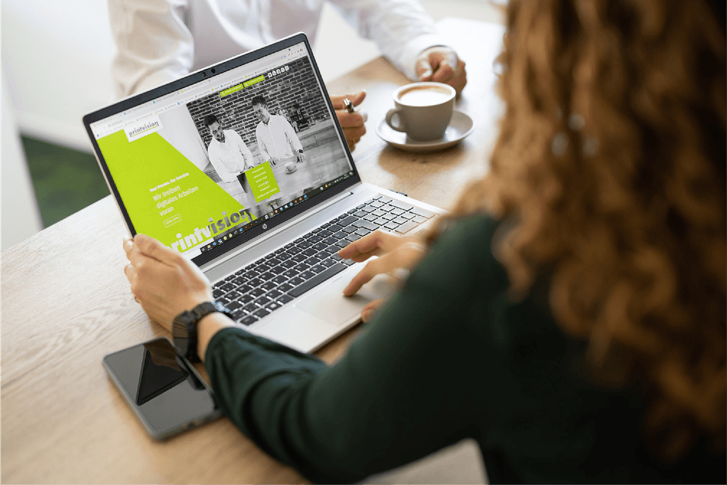 Eine Frau sitzt an einem Tisch im Büro und schaut sich auf dem Laptop eine Website der Firma printvison an, gegenüber sitzt ein Mann der gerade einen Cappucino trinkt