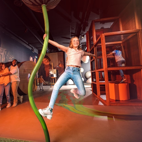 Una joven niña se balancea alegremente en un gran poste verde en un parque infantil cubierto, rodeada por otros niños y adultos.