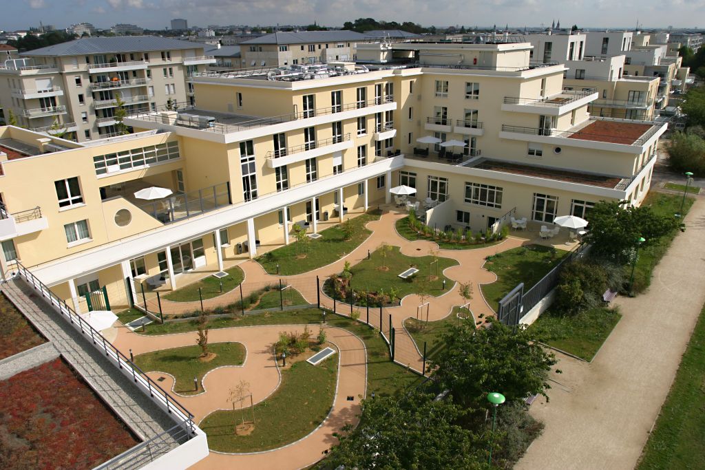 Aerial view of residential facility with cream-colored building, interior courtyard gardens, green landscaping, and surrounding area