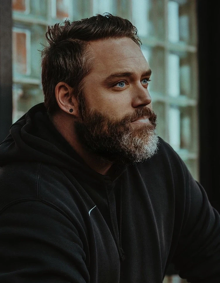 A man with a thick beard and short hair looks thoughtful while sitting indoors. He's wearing a black hoodie. Soft lighting highlights his contemplative expression.
