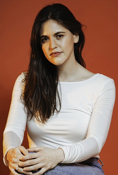 A woman with long, dark brown hair sits against a solid orange-brown background. She wears a white long-sleeved shirt and blue jeans. Her hands are clasped in front of her, and her gaze is directed slightly to her left. She has a neutral or slightly serious expression.