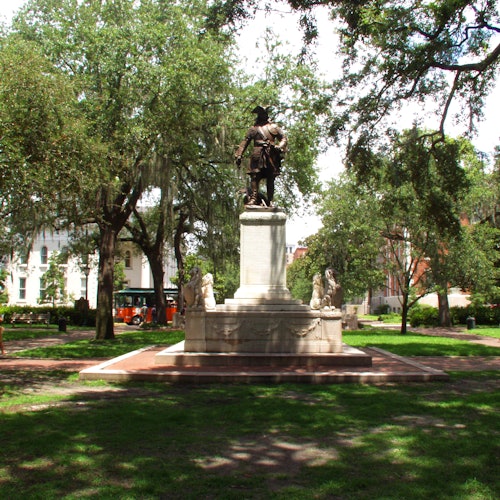 Статуя исторической фигуры стоит на пьедестале в зеленом парке, окружённом деревьями и зданиями на заднем плане.
