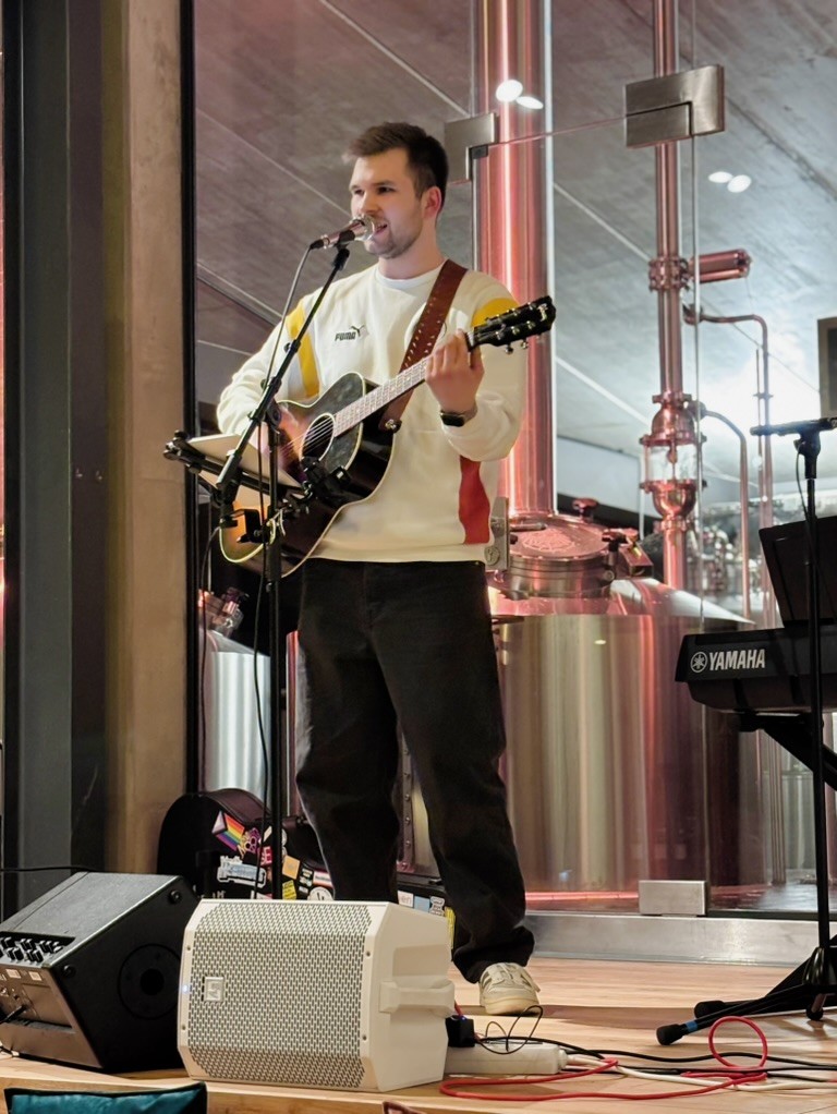 Ein Musiker mit weißem Pullover und Gitarre steht auf einer kleinen Bühne in einem Raum mit hohen Fenstern und industriellem Flair.