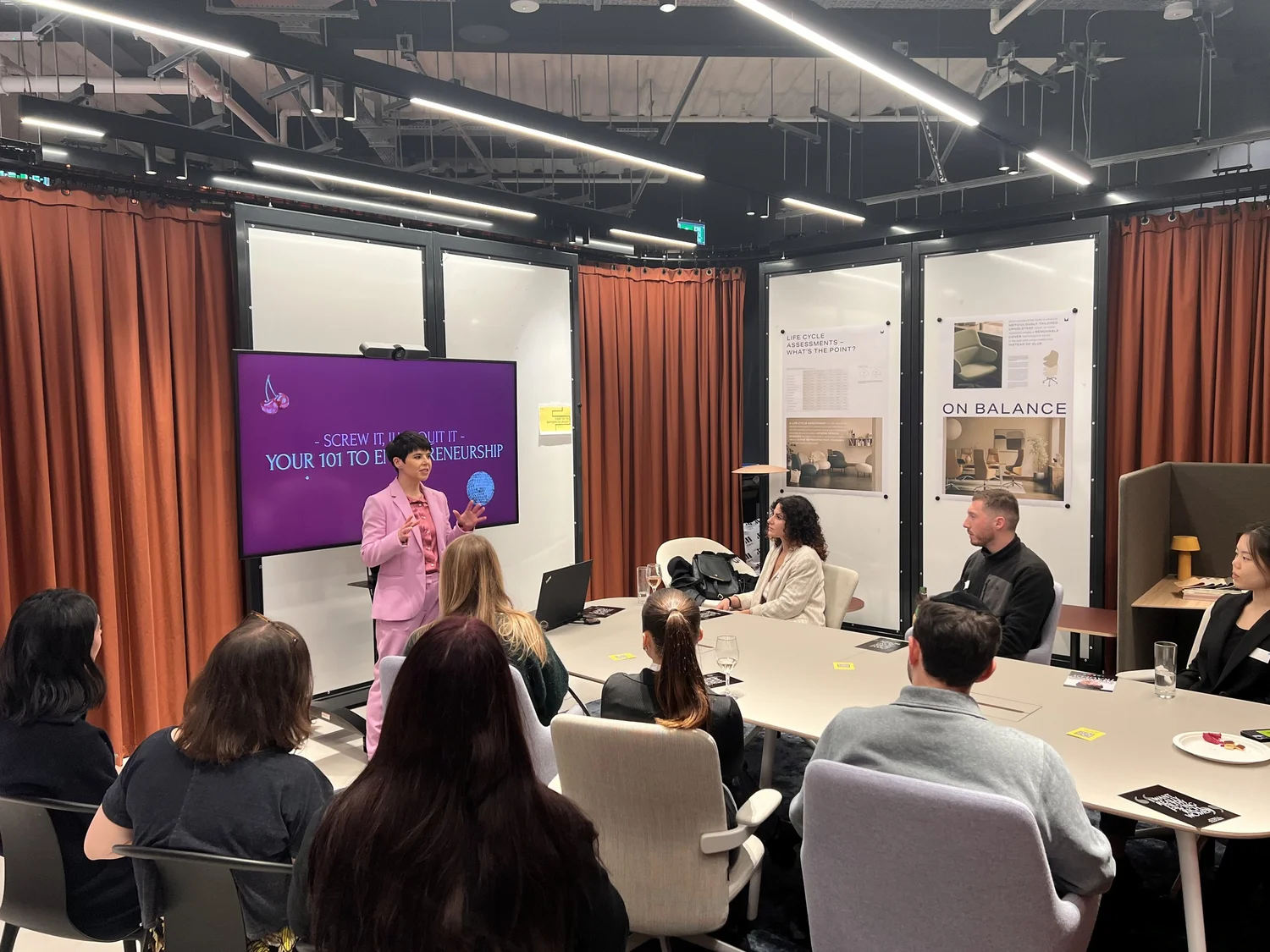 Group of people sitting at a long table looking at a speaker in a pink suit
