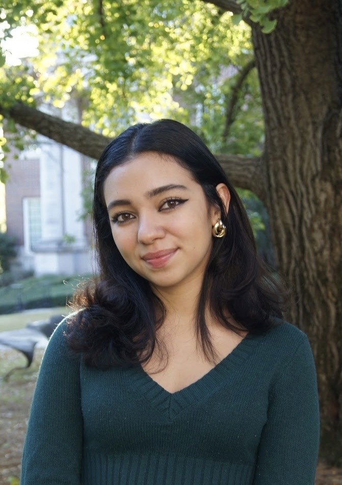A person stands outdoors in a natural setting with lush green trees under a clear sky, wearing a dark sweatshirt, and looking into the distance.