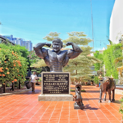 Statue of a muscular man flexing, with a plaque below, surrounded by plants and a red-tiled walkway. Small animal sculptures nearby.