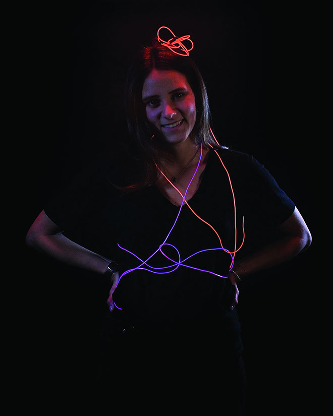 A woman with long dark hair stands against a black background. She wears a black top and a necklace with a pendant. Around her neck are glowing lines in red and purple, resembling a neon sign. Her hands are on her hips, and she looks directly at the camera with a slight smile.