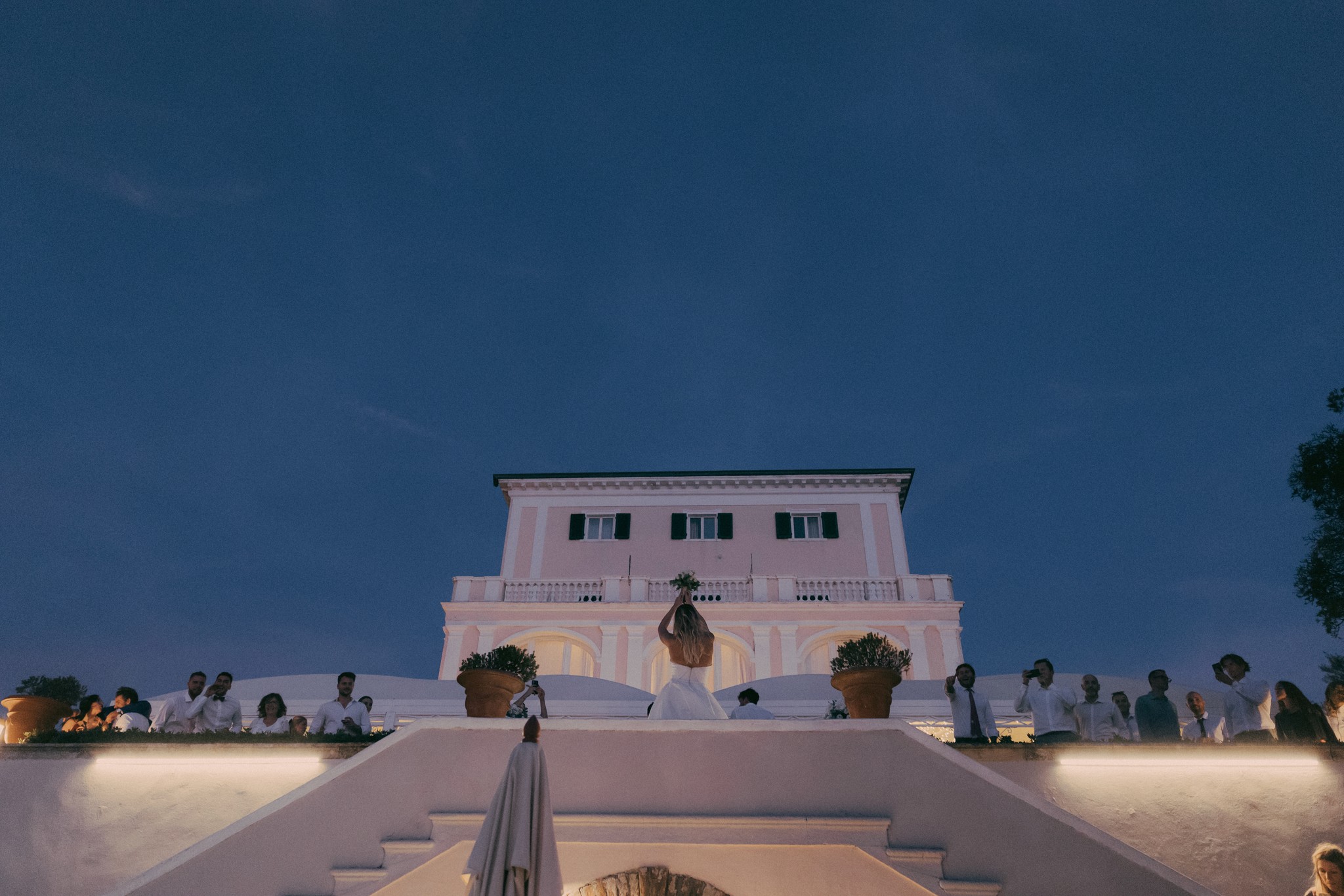 Sposi e ospiti sulla terrazza illuminata di sera a Bolgheri, reportage di matrimonio al tramonto con cielo blu
